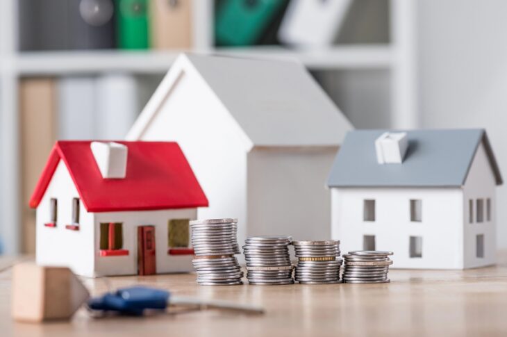 Miniature house models behind stacks of coins on a wooden table, representing real estate and investment
